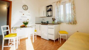 Cocina compacta con armarios blancos, una pequeña mesa de comedor con sillas amarillas, un reloj en la pared y una ventana con cortinas florales.