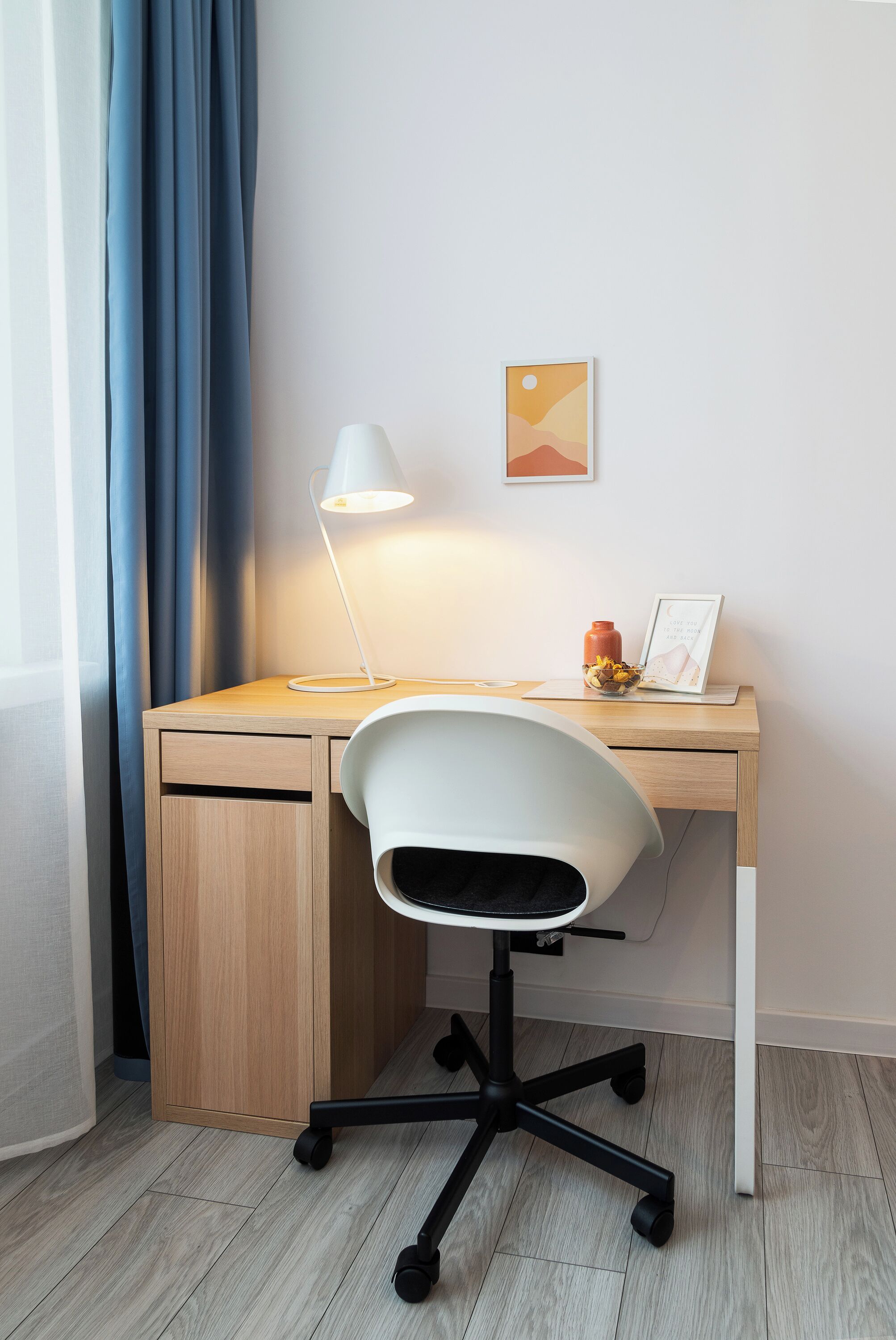 A modern workspace with a wooden desk, a white office chair, a desk lamp, a framed picture, and a small bowl of fruit.