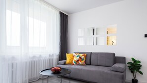 A modern living room with a grey sofa, a round coffee table with a bowl of fruit, sheer curtains, and a potted plant. - Family - by Kairos Apartments (Gliwice)