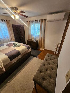 Un dormitorio con cama, mesita de noche, banco, ventilador de techo y ventana con cortinas.