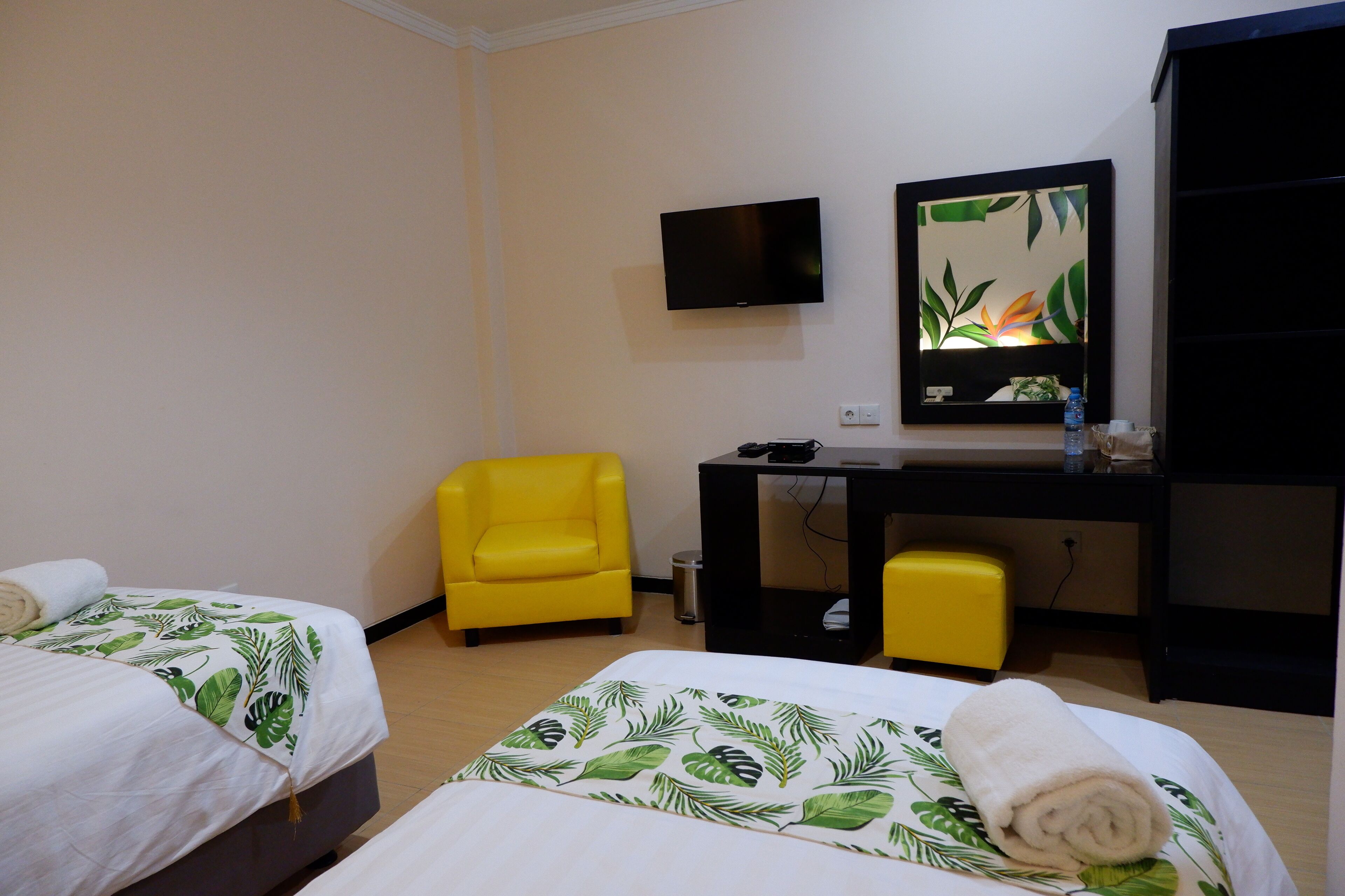 Habitación de hotel con dos camas, una silla amarilla, un escritorio con televisor y un espejo.