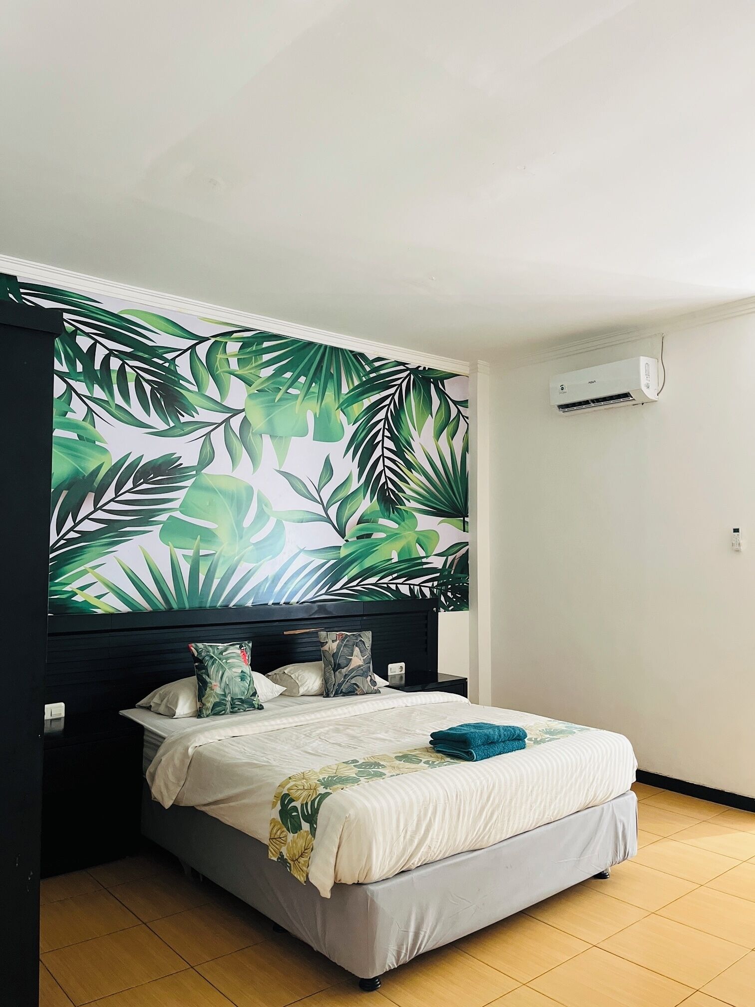 A bedroom with a bed, a headboard with a tropical leaf mural, an air conditioning unit on the wall, and a tiled floor.