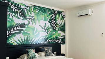 A bedroom with a bed, a headboard with a tropical leaf mural, an air conditioning unit on the wall, and a tiled floor.