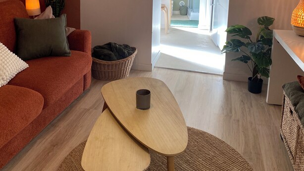 Una sala de estar acogedora con un sofá, una mesa de centro y una lámpara. Hay una planta en el suelo y otra en una mesa. Se ve un reloj en la pared.