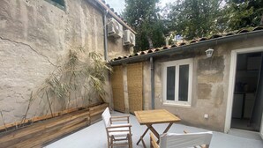 Un patio con una mesa y sillas de madera, una planta de bambú y un edificio con tejado de tejas y una ventana.