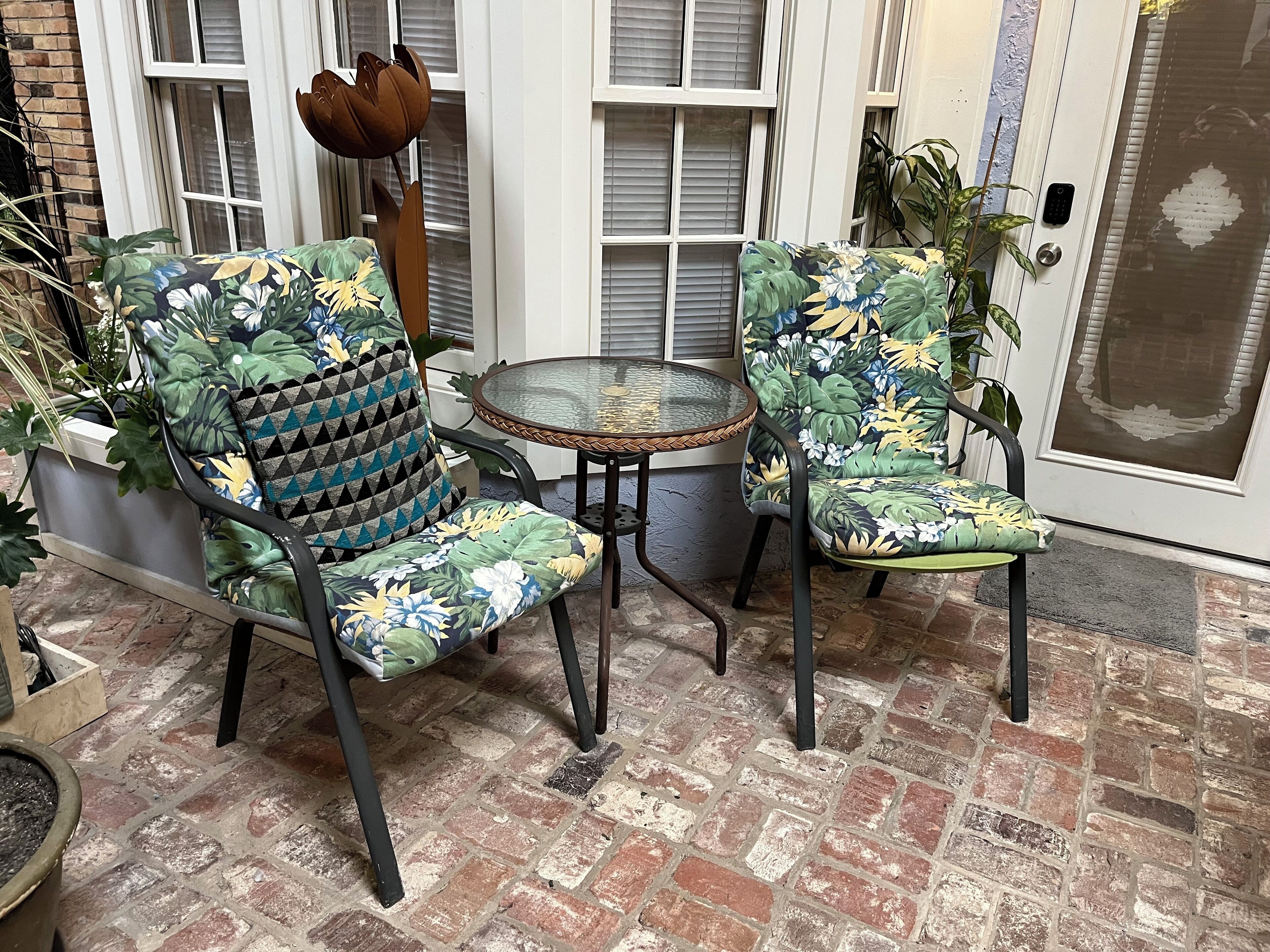 Deux chaises recouvertes d’un tissu à motifs floraux et une petite table ronde dont le bord est orné d’un motif tressé, disposées sur une terrasse en briques.
