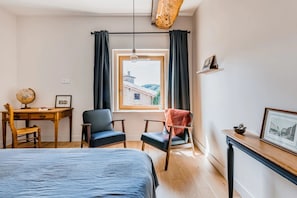 Un dormitorio con cama, dos sillas, un escritorio con un globo terráqueo, una ventana con cortinas y vistas a un edificio.