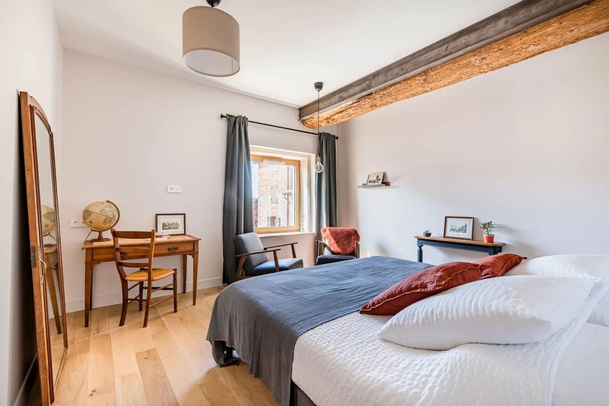 A bedroom with a wooden desk, a globe, a chair, a bed with white and red pillows, and a window with curtains.