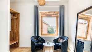 Een kamer met een groot raam, twee stoelen, een klein tafeltje en een houten garderobe.