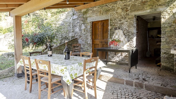 Une terrasse extérieure avec un mur en pierre, une table et des chaises en bois, un vase de fleurs et une porte rustique.