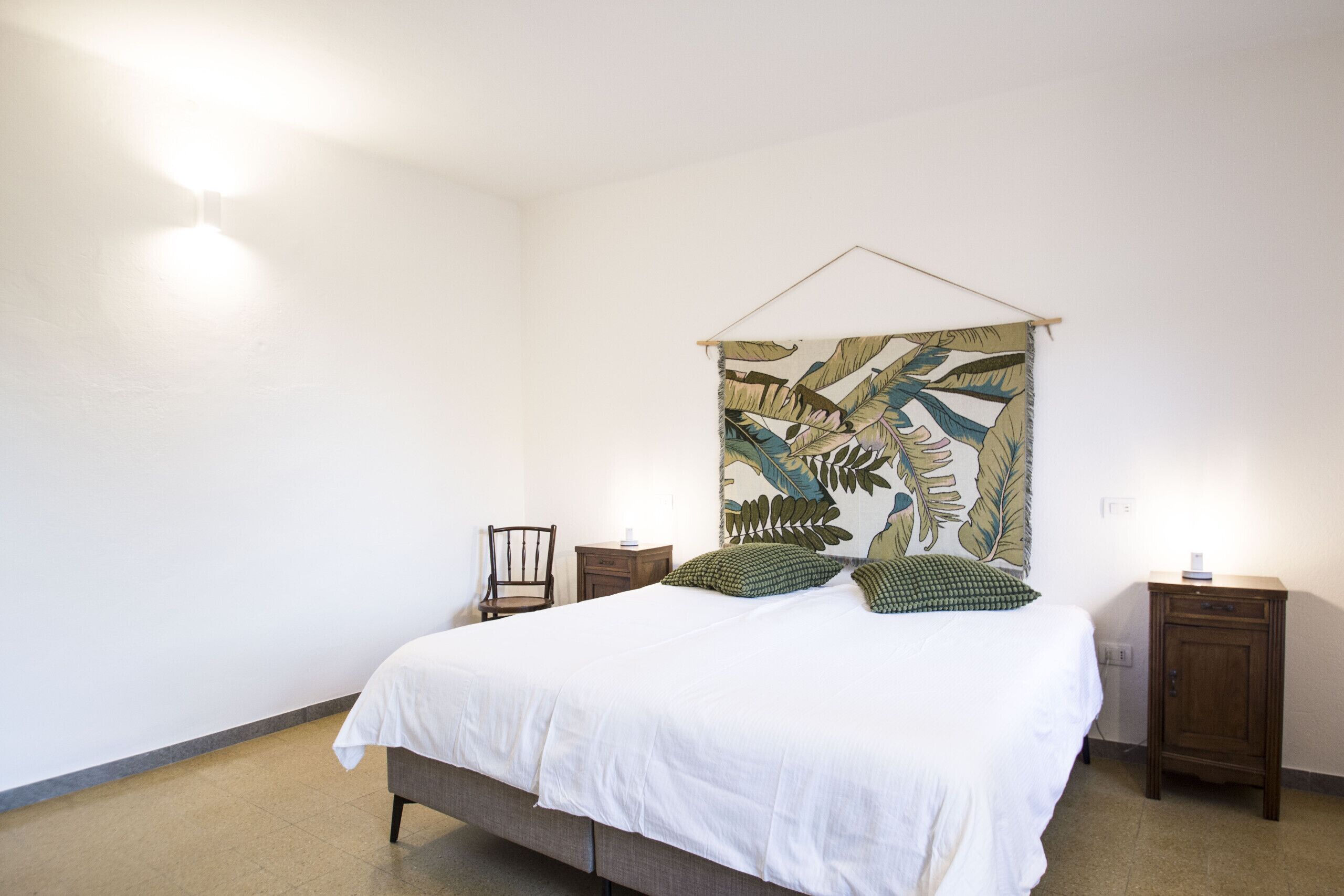 A bedroom with a bed, a hanging tapestry, two nightstands, and a chair.