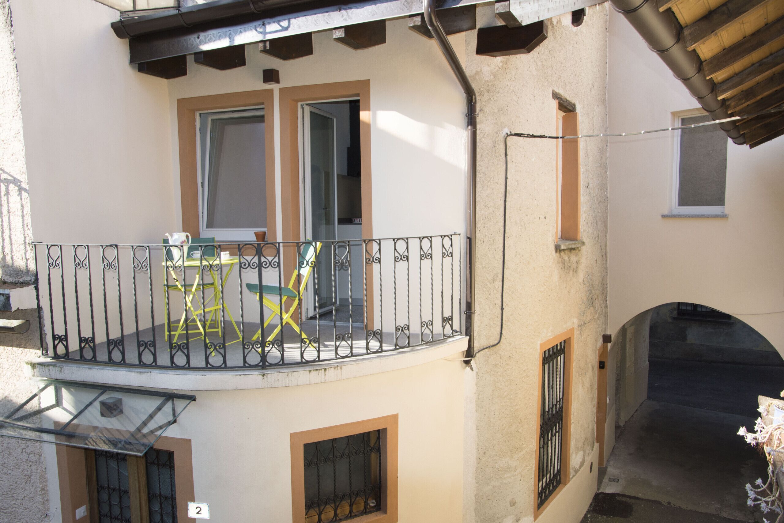 Un balcon doté d’une rambarde en métal, d’une petite table et de deux chaises. On y trouve deux portes et une fenêtre.