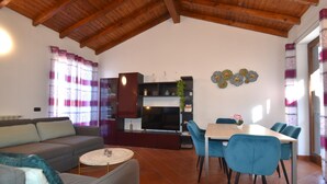 A living room with a sofa, a TV, a dining table with chairs, and a kitchen area with a TV and shelves.