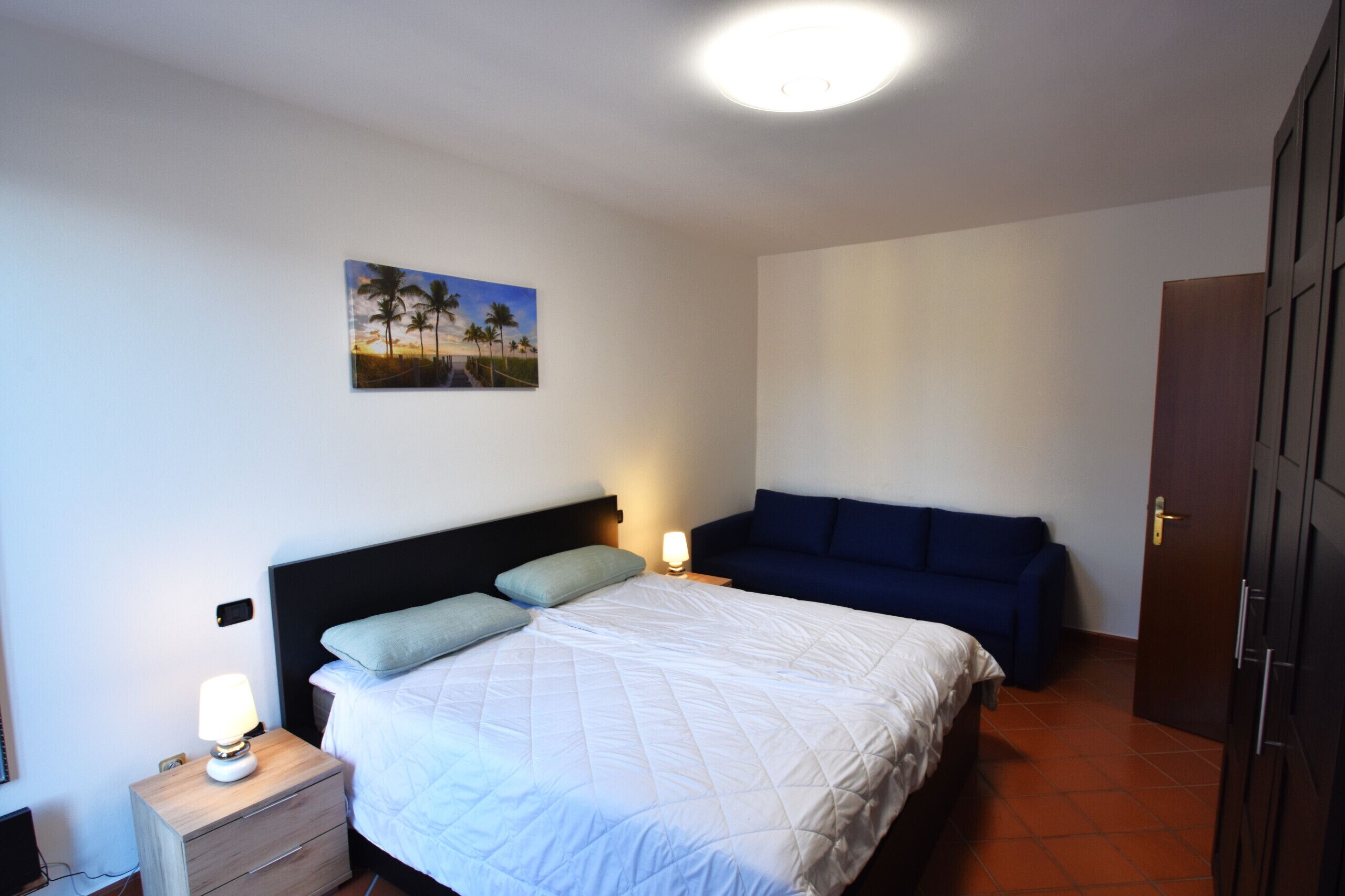 A bedroom with a bed, bedside table, nightstand, lamp, and a painting on the wall.