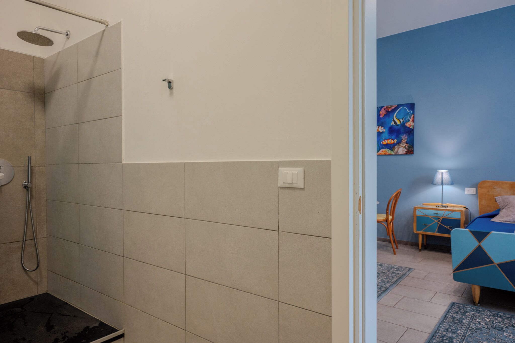 A bathroom with a shower, a toilet, and a sink. There is a bed with a blue headboard and a nightstand with a lamp.