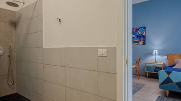 A bathroom with a shower, a toilet, and a sink. There is a bed with a blue headboard and a nightstand with a lamp.