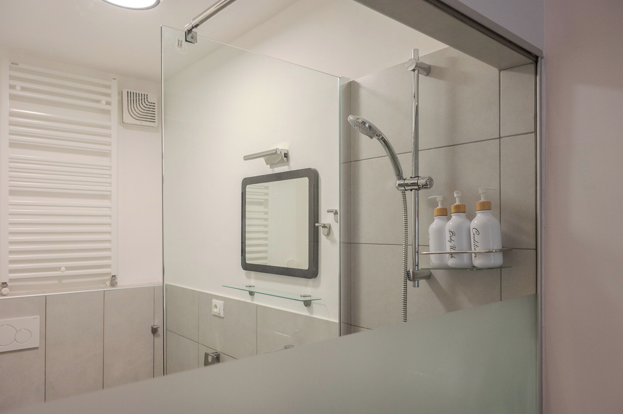 A modern bathroom with a glass shower enclosure, a wall-mounted mirror, and a shelf with toiletries.