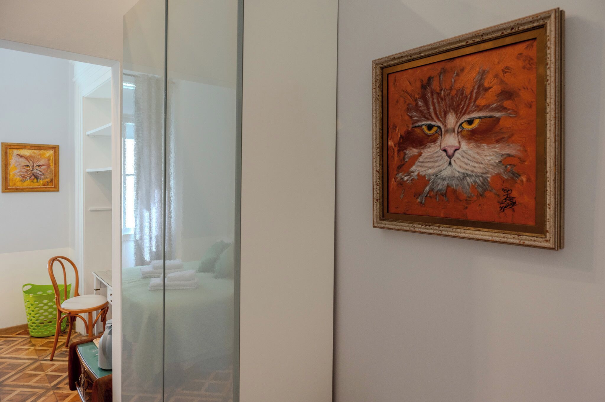 A room with a bed, a chair, a green trash bin, and a painting of a cat on the wall.