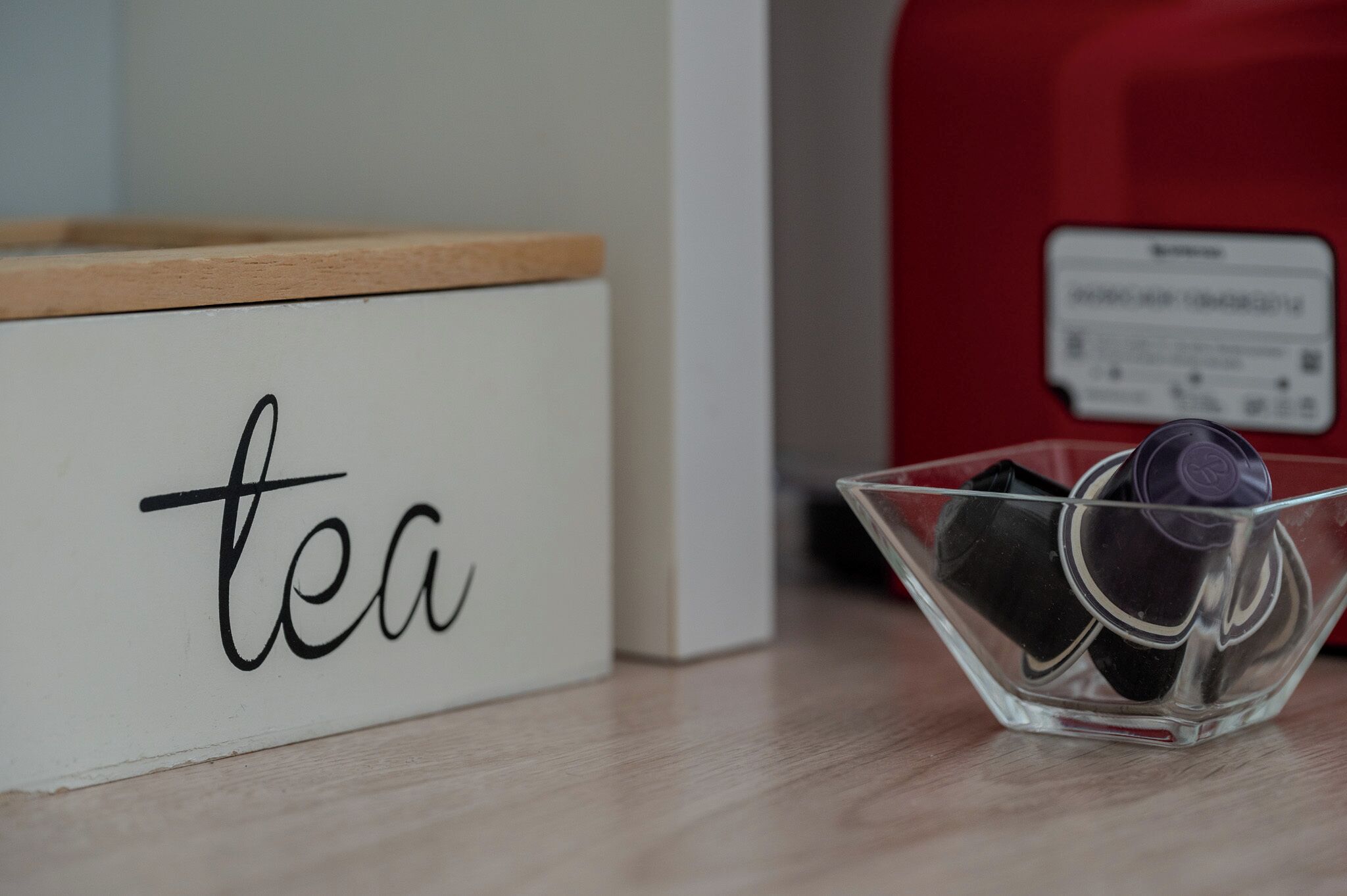 Eine weiße Box mit dem Wort 'tea', eine transparente Glasschale mit Teebeuteln und ein roter Behälter im Hintergrund.