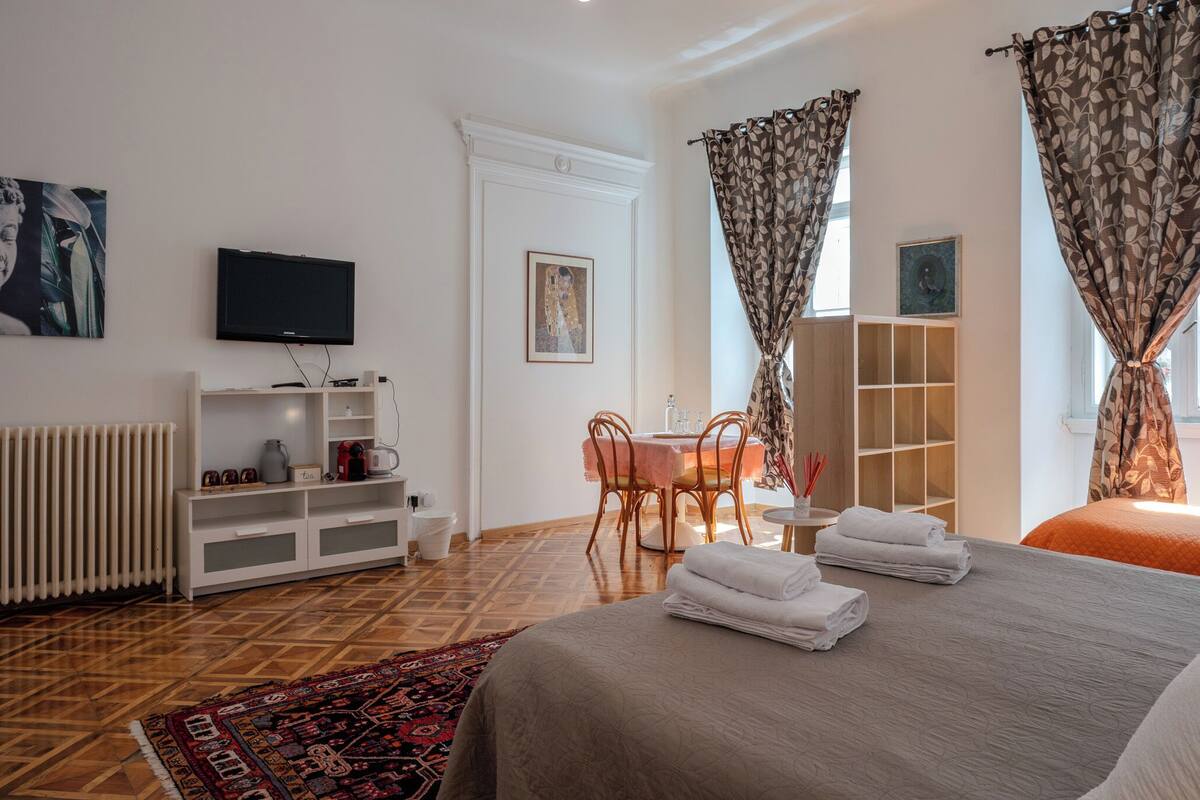 Ein Hotelzimmer mit Bett, Fernseher, Esstisch mit Stühlen, Heizkörper und Vorhängen.