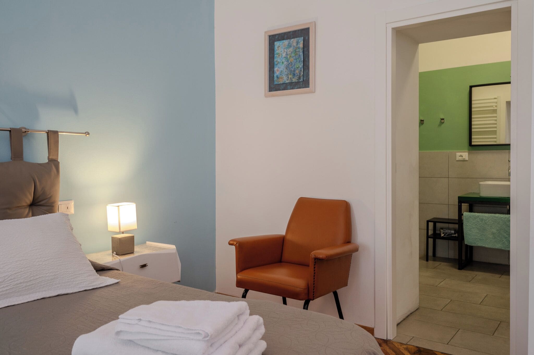 A hotel room with a bed, a chair, a nightstand with a lamp, and a small table with a towel.