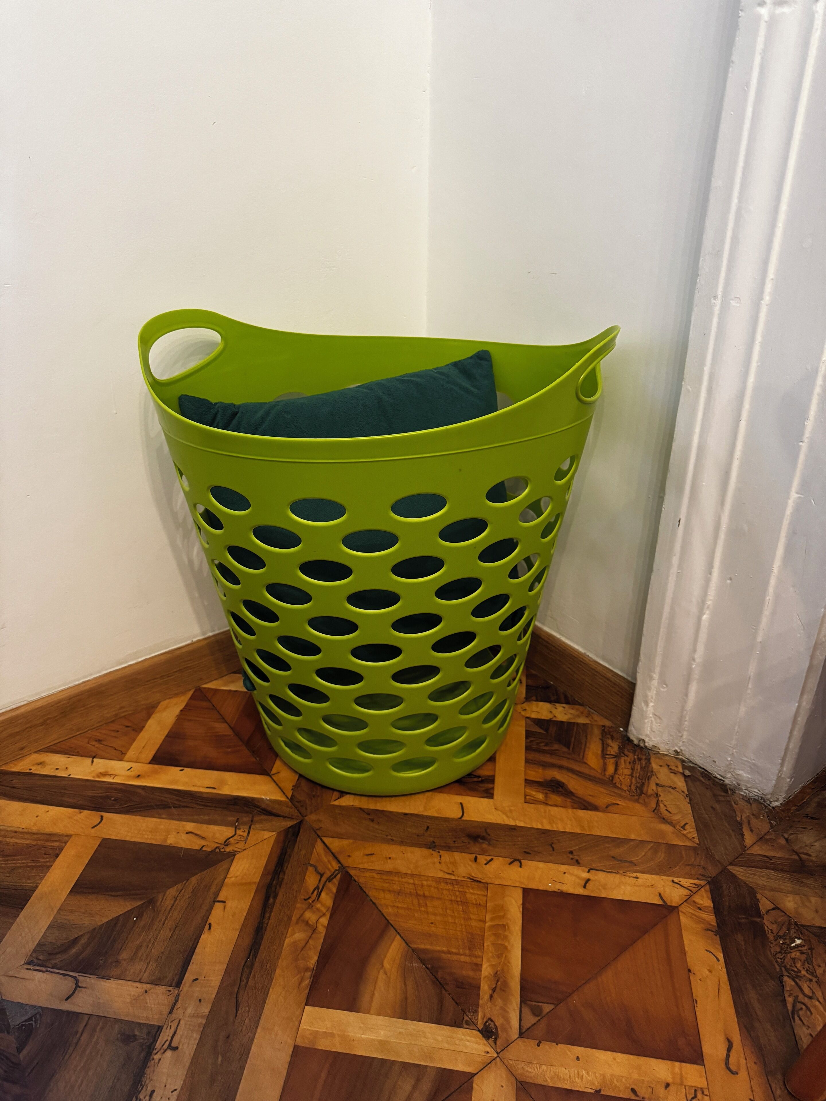 A green laundry basket with a cushion inside, placed on a wooden floor with a geometric pattern.