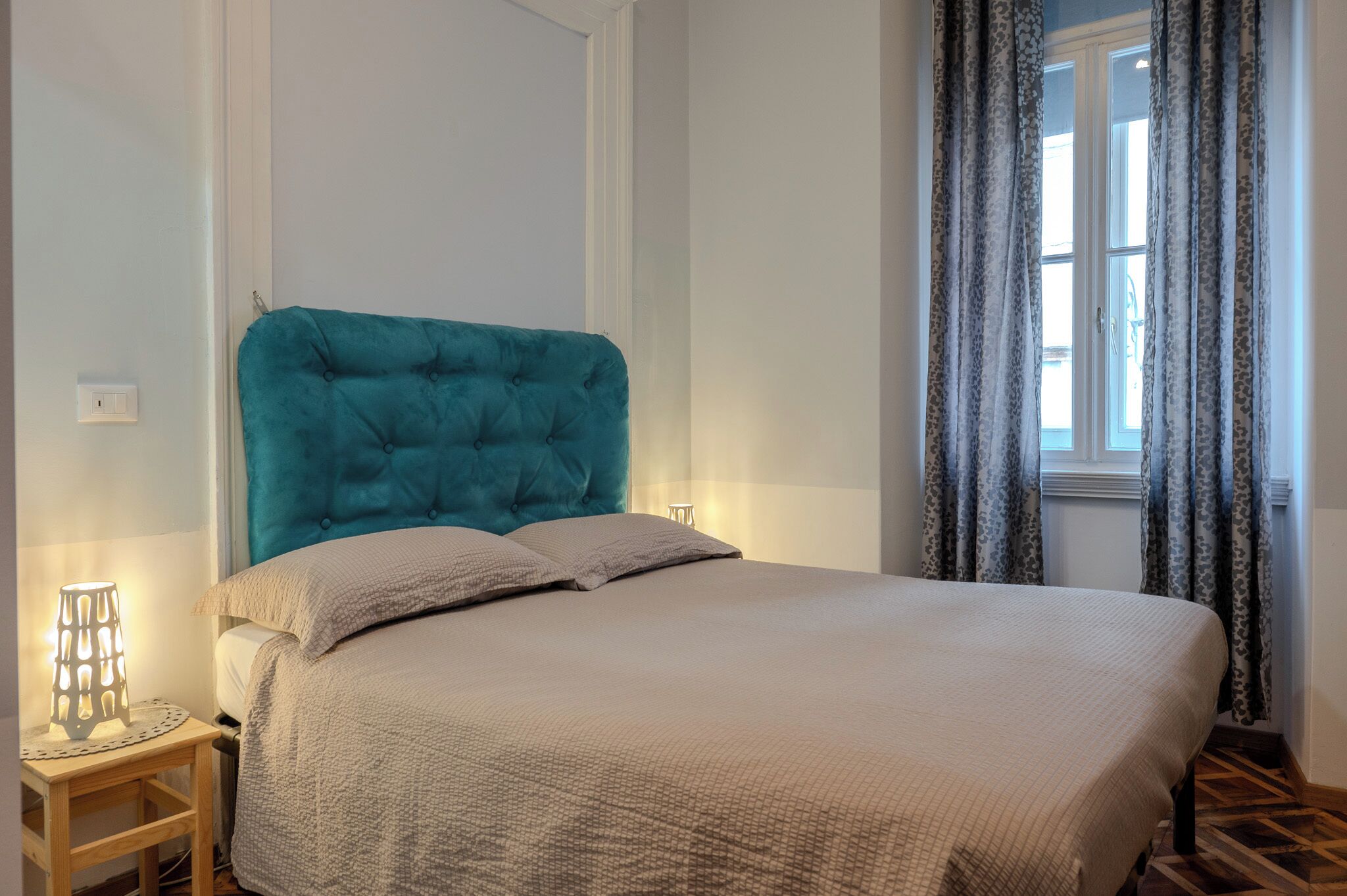 A bedroom with a tufted teal headboard, a beige bedspread, and a bedside table with a lamp.