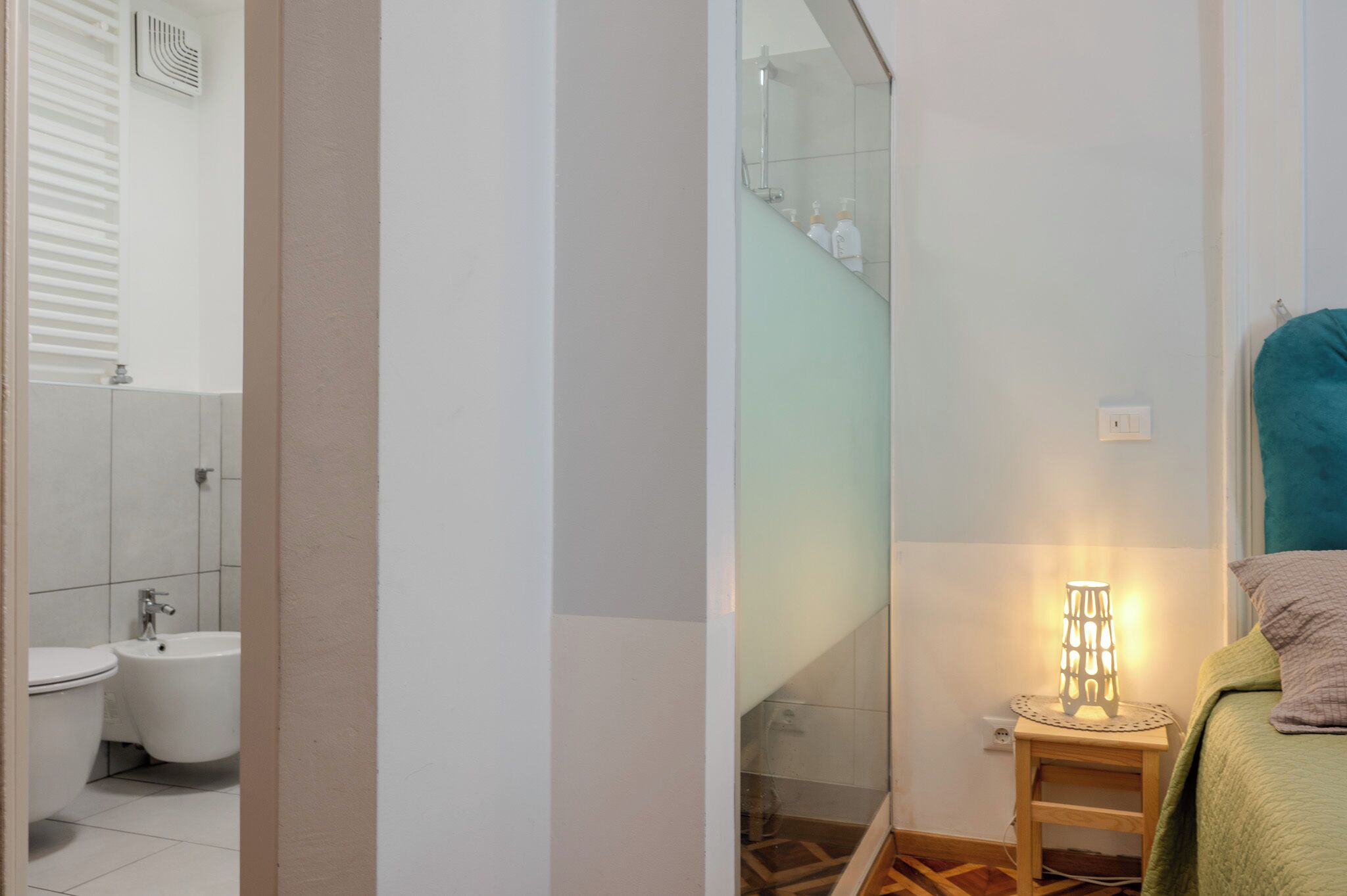 Ein Badezimmer mit Toilette, Bidet und Dusche. Ein kleines Nachttische mit Lampe steht neben einem Bett.