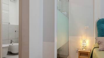 Ein Badezimmer mit Toilette, Bidet und Dusche. Ein kleines Nachttische mit Lampe steht neben einem Bett.