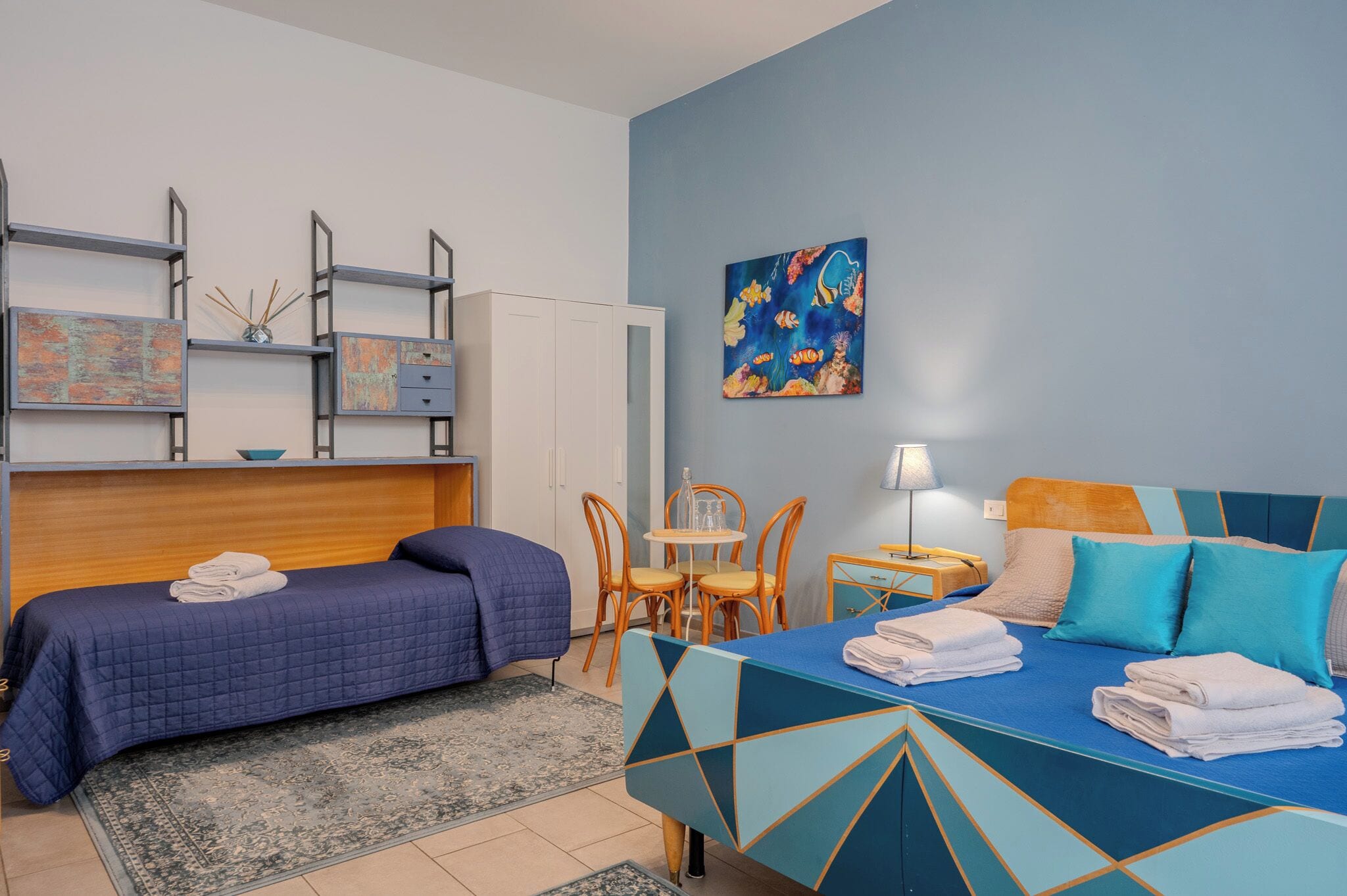A hotel room with two beds, a dining table, chairs, a small table with a lamp, and artwork on the wall.