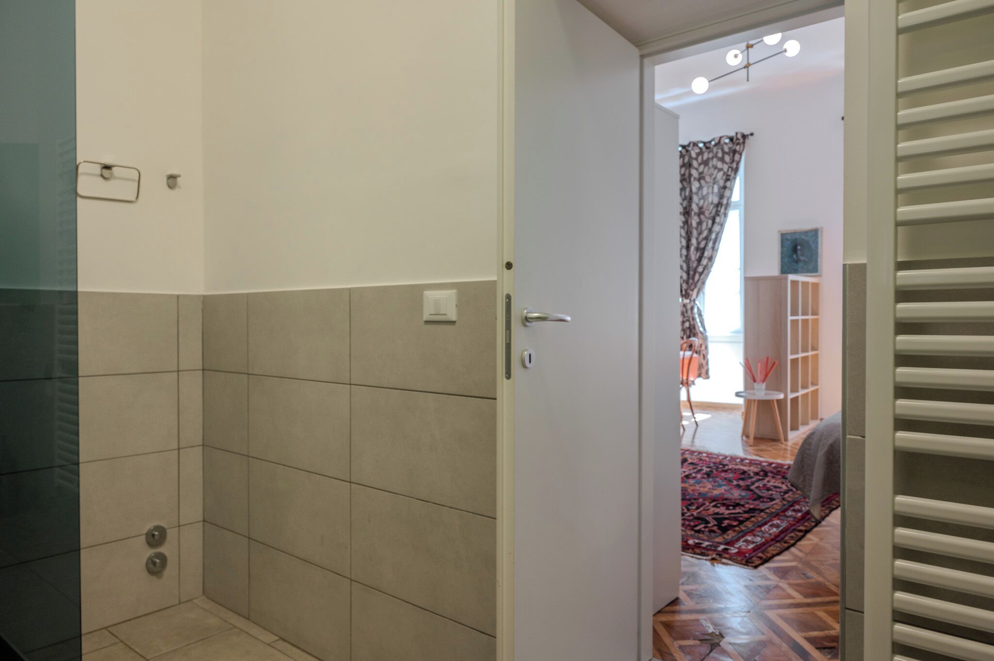 A bathroom with a shower, a toilet, and a sink. There is a door leading to a living area with a rug and a small table.