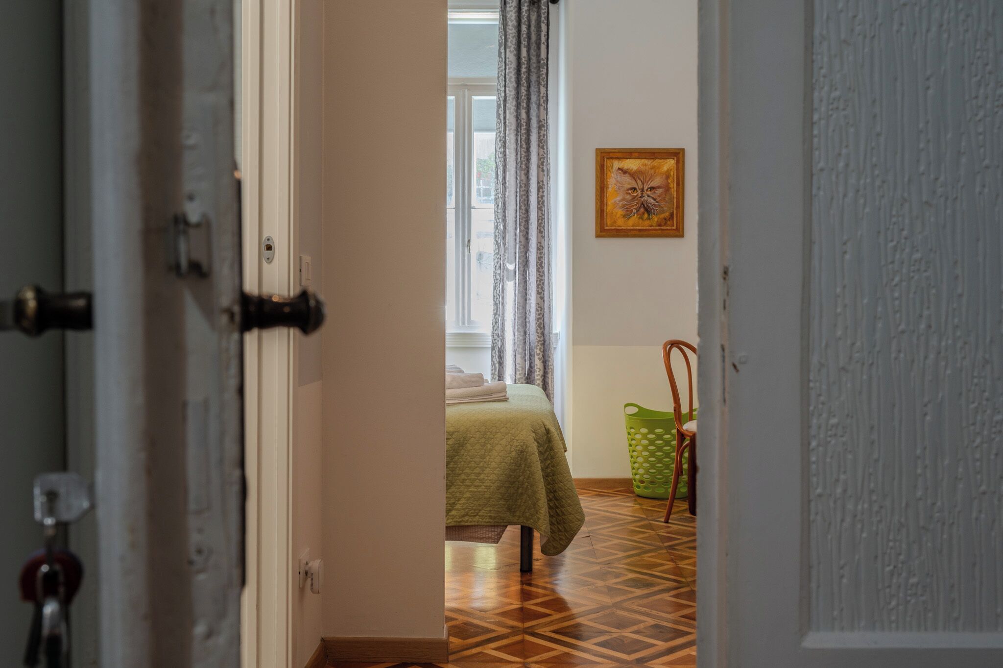 An open door leading to a room with a bed, a window with curtains, a green chair, and a wall-mounted painting.