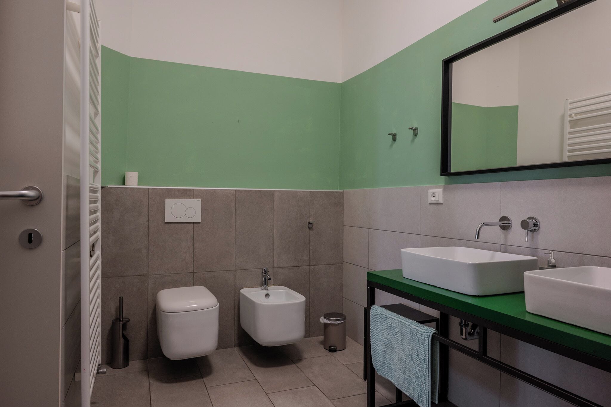 A bathroom with a green accent wall, a white toilet, a bidet, a sink with a mirror, and a towel rack.