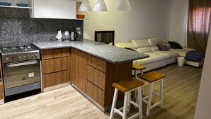 Une cuisine moderne dotée d’une cuisinière, d’un four et d’un plan de travail en granit intégrés. On y trouve un îlot central en bois avec des tabourets, un canapé blanc et un plafond en bois.