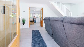 A modern living room with a grey sofa, a wooden door, a staircase, and a kitchen in the background. - Erin's Cottage in Merthyr Tydfil (Merthyr Tydfil)