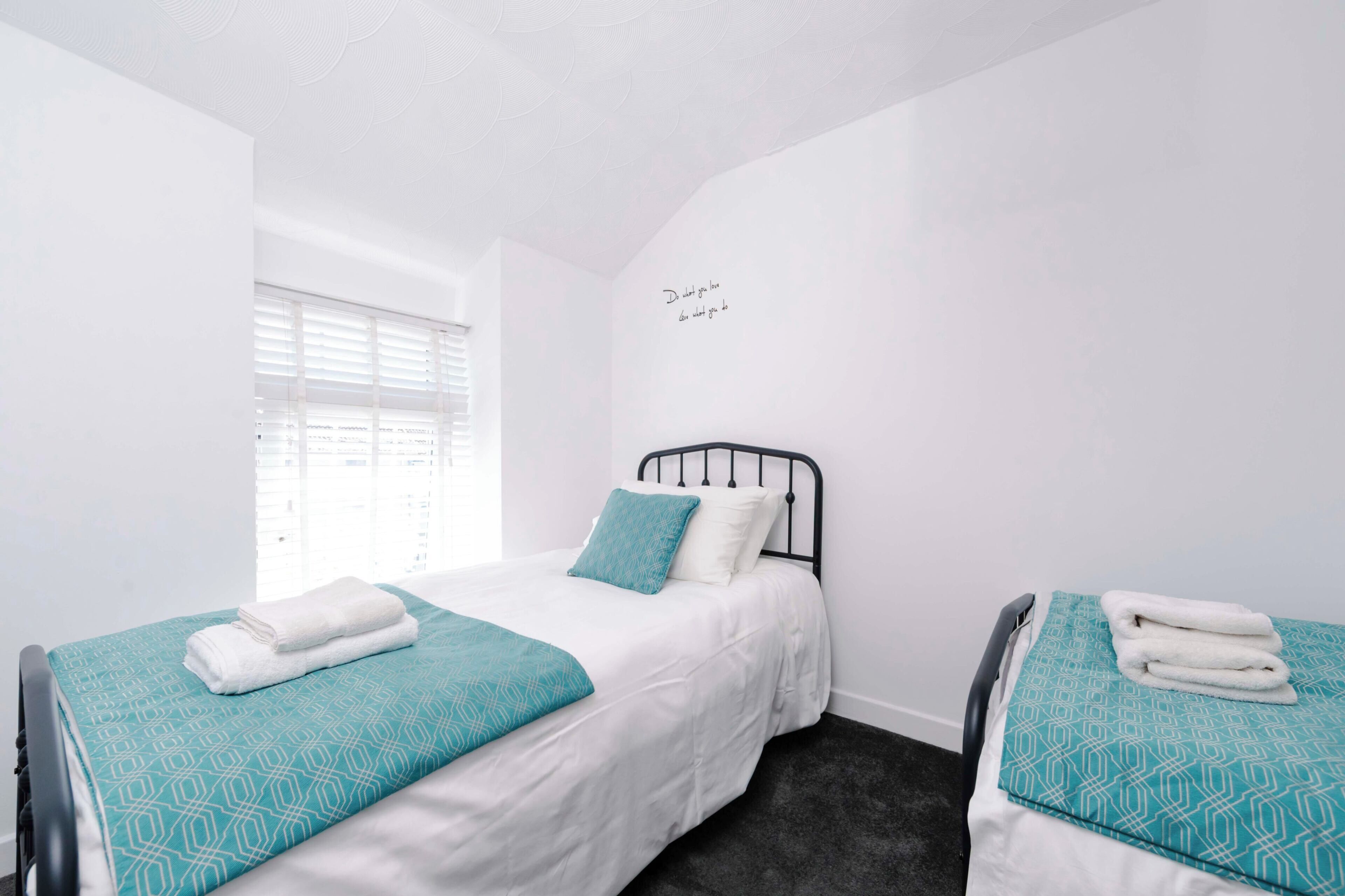 A bedroom with two beds, each with a teal patterned bedspread and white linens, a window with blinds, and a wall with a motivational quote.