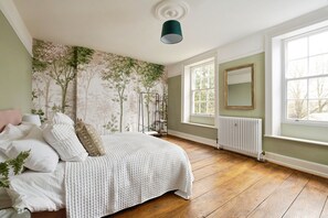 Une chambre à coucher avec un lit, un radiateur, une fenêtre et un mur recouvert d’un papier peint à motifs floraux.