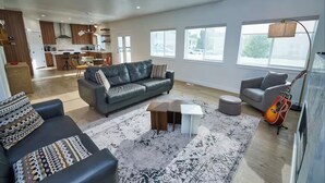 A spacious living room with a grey sofa, a smaller grey armchair, and a coffee table. There is a guitar leaning against the wall and a lamp on a side table. - 3 Bedroom, 4 bed/3 Bath, Close to Mountains, Downtown and Airport (Midvale)