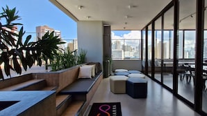 A modern rooftop lounge with seating areas, large windows offering city views, and a clear sky. - COBAR0131 - Edifício Único Barra (Salvador)
