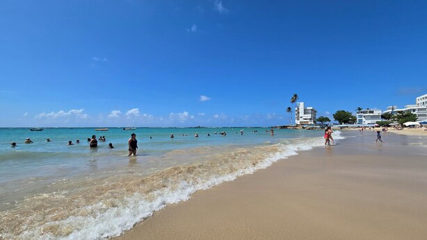 Une plage avec une eau d’un bleu limpide, des gens qui se baignent et qui se promènent sur le sable, et des bâtiments en arrière-plan.