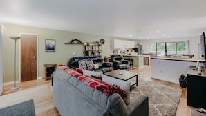 A modern living room with a grey sofa, coffee table, and a red and white blanket. There is a kitchen area with white cabinets and a large window. - 52mbr - Hot Tub - Pets Ok - Generator - Wifi (Deming)