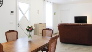 Een eethoek met een houten tafel en stoelen, een vaas met bloemen, een bruine leren bank, een televisie en een witte deur met een raam.