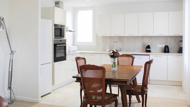Een moderne keuken met witte kasten, een eettafel met vier houten stoelen, een magnetron, een oven en een spoelbak.