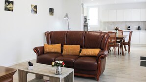 Een moderne woonkamer met een bruine leren bank, een houten salontafel en een vaas met bloemen. Er is een eethoek met een tafel en stoelen, en een keuken op de achtergrond.