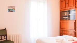 Een slaapkamer met een bed, een stoel, een radiator, een raam met gordijnen en een houten kast met boeken.