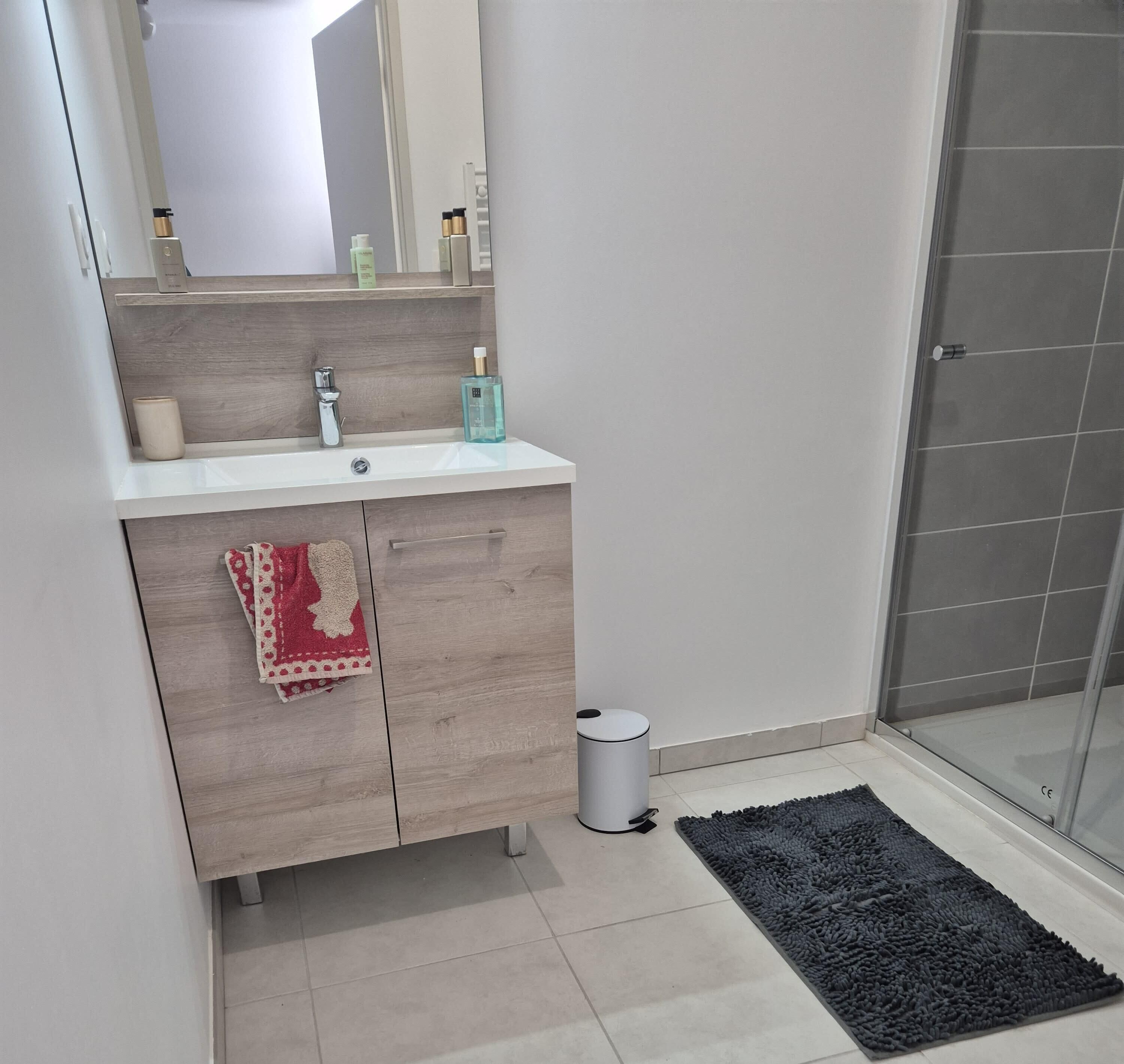 Une salle de bain avec un lavabo blanc, un miroir, une serviette, une poubelle et un espace douche.