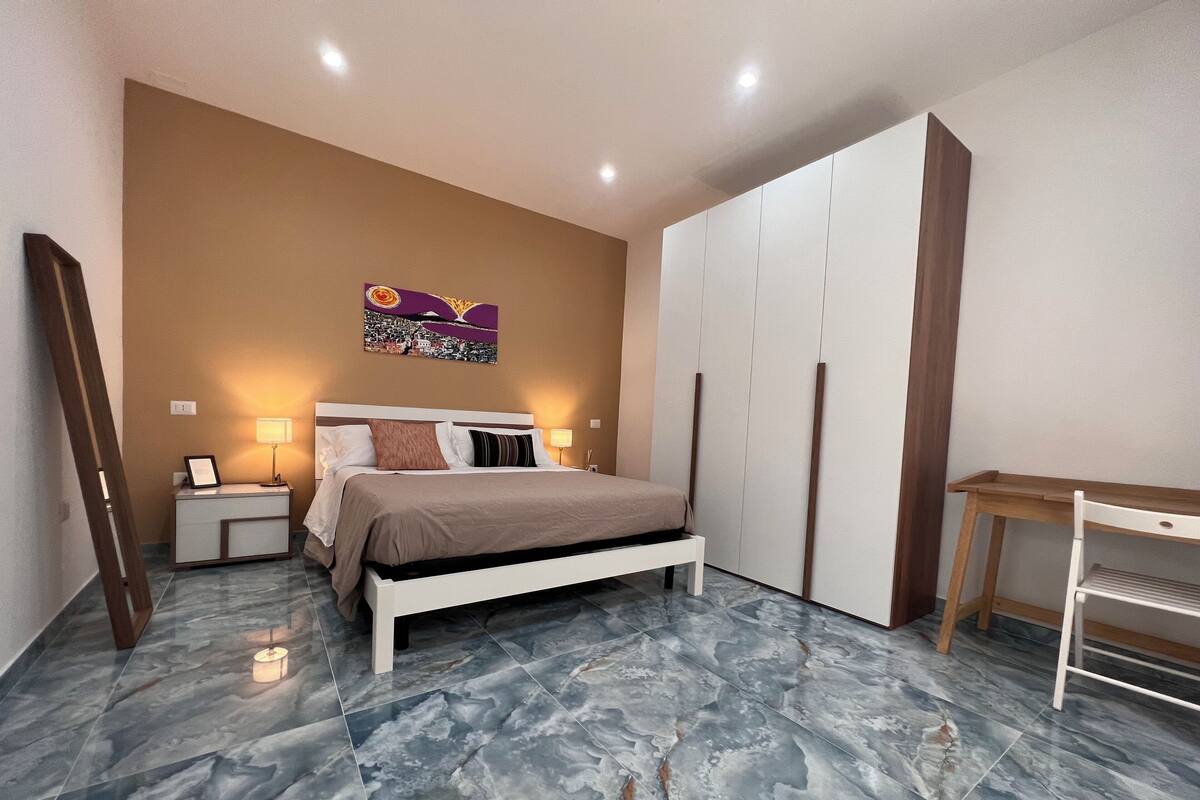 A bedroom with a bed, nightstands, a desk, a wardrobe, and a wall-mounted artwork.
