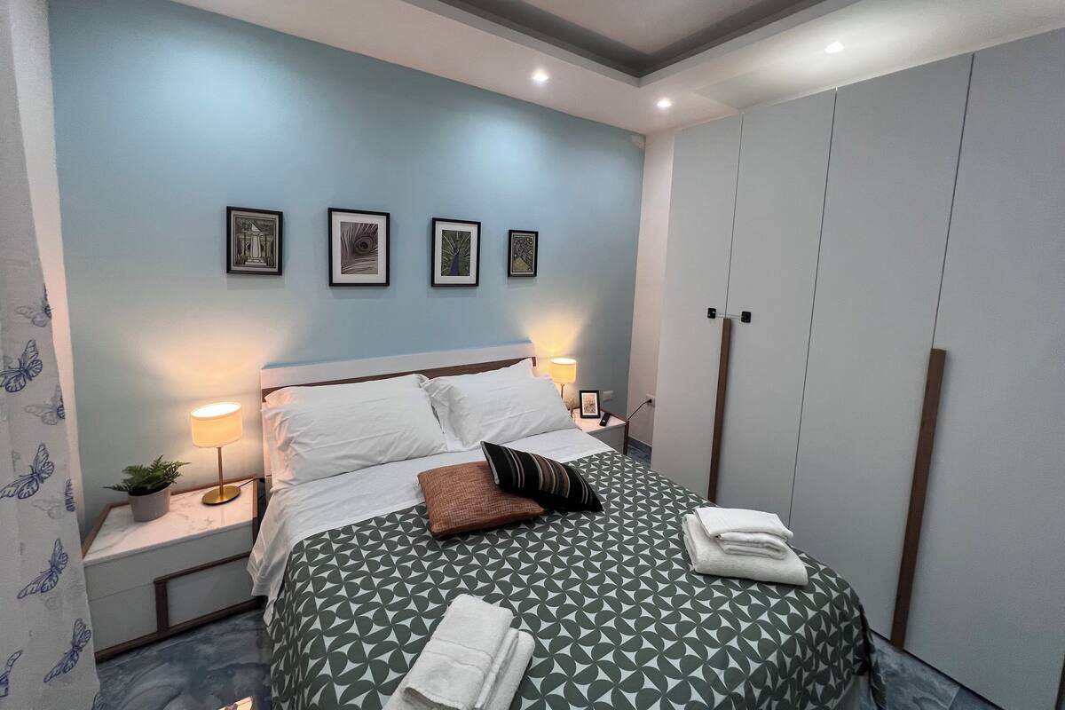 A modern bedroom with a geometric-patterned bedspread, a nightstand with a lamp, a wall-mounted shelf with framed pictures, and a wardrobe.