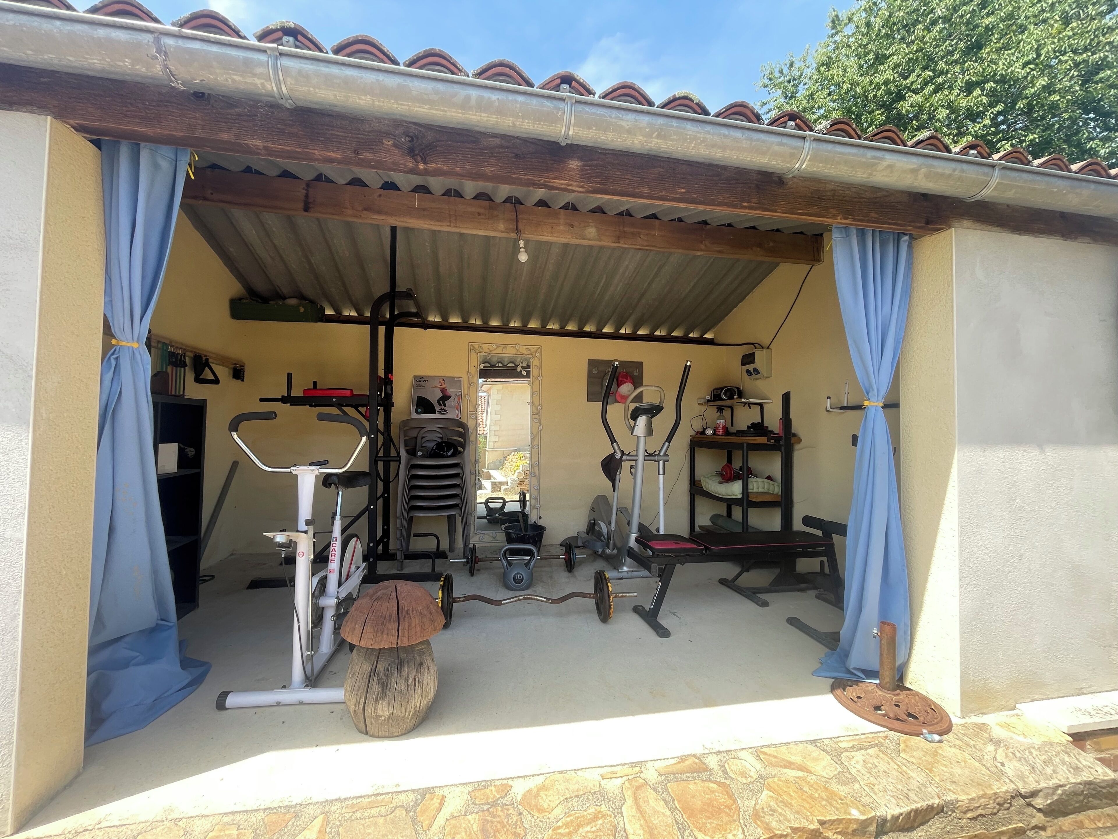 Un'area fitness all'aperto, riparata, con attrezzature per esercizi, tra cui una cyclette, un ellittico e pesi.
