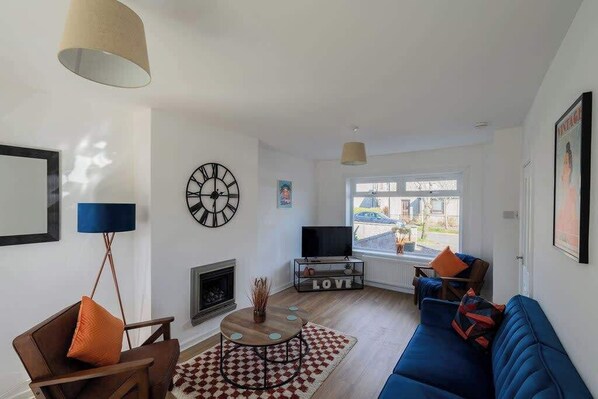 Una sala de estar con sofá, sillón, televisión, chimenea y un ventanal grande.